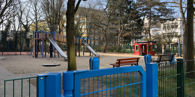 Spielplatz mit zwei Rutschen, einer großen Sandfläche gerahmt von Bäumen