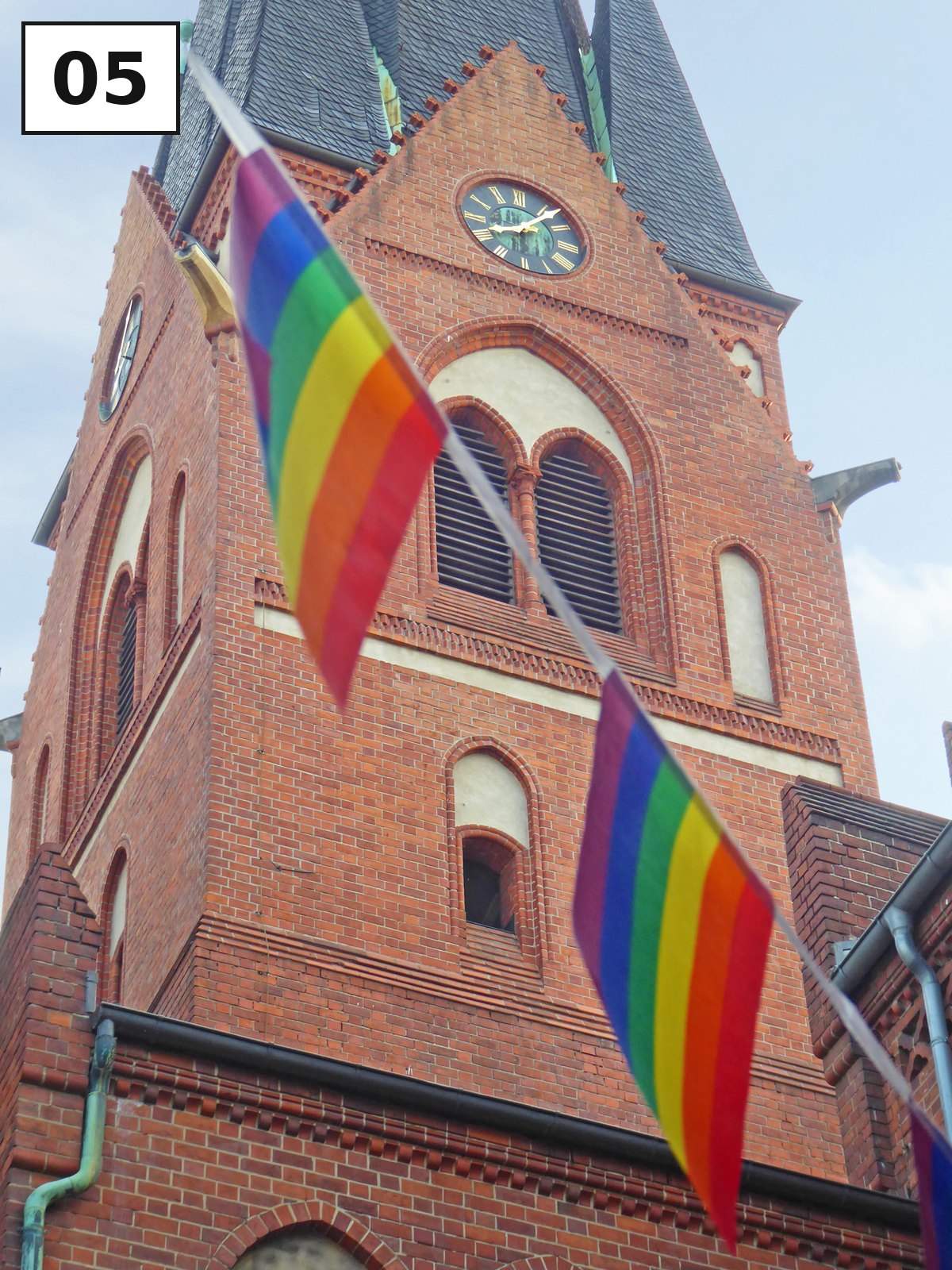 Backsteinkirche mit Turmuhr, zwei Regenbogenfahnen wehen am Turm